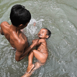 A father bathes his child while teaching him how to swim. He hopes that his children will become fishers in the Khone Falls.